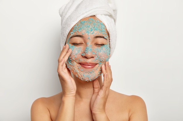 Joven alegre aplica exfoliante natural en la cara, toca las mejillas, mantiene los ojos cerrados, usa una toalla, se somete a procedimientos de belleza después de la ducha, modela en interiores. Modelo femenino con sal marina azul en la piel