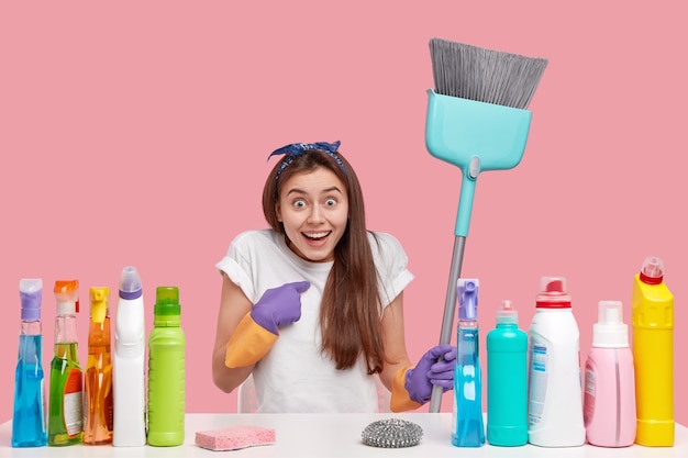 Foto gratuita joven alegre asombrada se indica a sí misma, trabaja en el servicio de limpieza, sostiene una escoba, se sienta en el escritorio con detergentes y desodorante