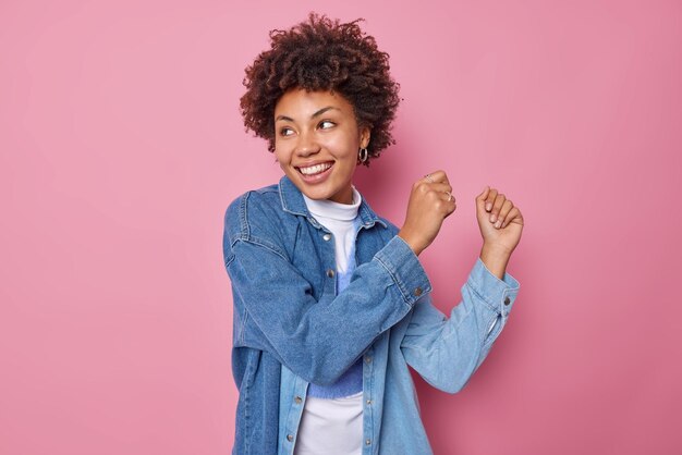 Una joven alegre baila movimientos despreocupados activamente vestida con una camisa de mezclilla sonríe ampliamente tiene un estado de ánimo optimista enfocado lejos aislado sobre un fondo rosado disfruta de su lista de reproducción favorita hace bailar la victoria