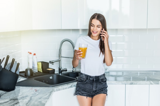 Joven alegre bebiendo jugo de naranja mientras habla por teléfono móvil y de pie cerca de una mesa de la cocina