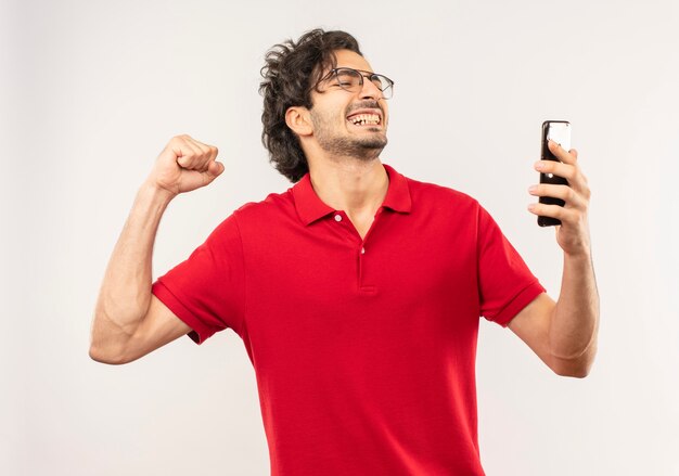 Joven alegre en camisa roja con gafas ópticas mira el teléfono y levanta el puño aislado en la pared blanca