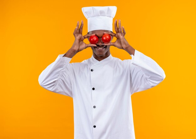 Joven alegre cocinero afroamericano en uniforme de chef cierra los ojos sosteniendo tomates con las manos aisladas en la pared naranja