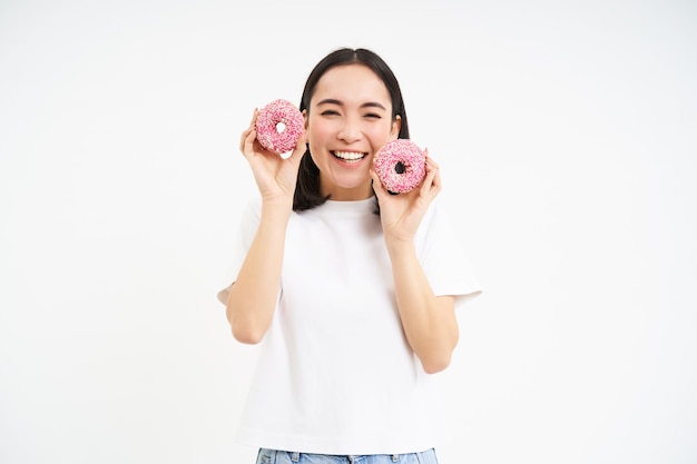 Foto gratuita una joven alegre comiendo dos donuts rosados glaseados sobre fondo blanco.