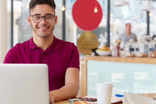 Foto gratuita un joven alegre y complacido conversa en línea con amigos del extranjero, se sienta frente a una computadora portátil, está conectado a internet 4g en la cafetería, usa lentes ópticos para una buena visión, le gusta su trabajo