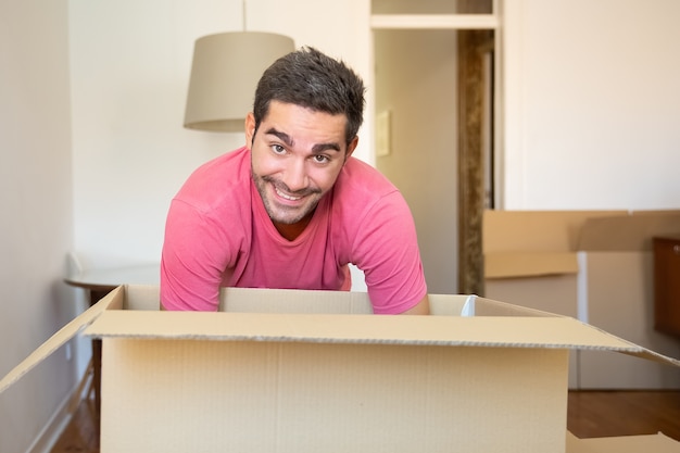 Joven alegre desembalaje de cosas en su nuevo apartamento, abriendo la caja de cartón,