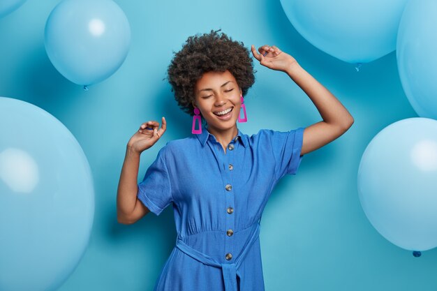 Joven alegre y elegante mujer afroamericana baila con las manos levantadas, disfruta celebrando la fiesta, viste un vestido azul de moda, se mueve