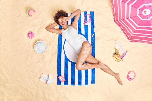 Una joven alegre esconde la cara con sonrisas en las manos con gusto usa poses de traje de baño blanco en una toalla a rayas toma el sol en la playa tiene un día perezoso viaja al extranjero para las vacaciones de verano Concepto de tiempo de vacaciones