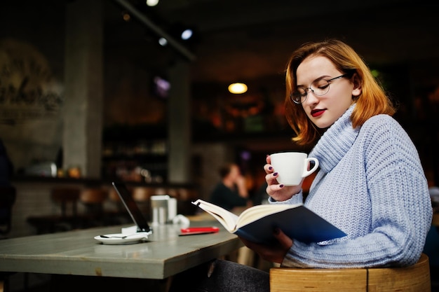 Una joven alegre y hermosa mujer pelirroja con anteojos usando el panel táctil de su teléfono y su cuaderno mientras se sienta en su lugar de trabajo en el café con una taza de café