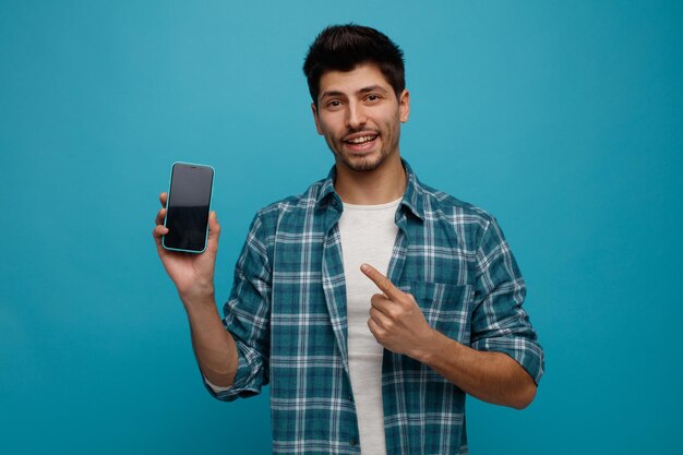 Joven alegre mostrando el teléfono móvil a la cámara apuntándolo mirando a la cámara aislada en el fondo azul