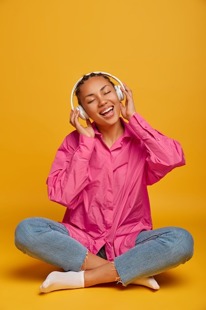 Joven alegre mujer étnica disfruta de la música en el piso, se sienta con las piernas cruzadas, usa camisa rosa, jeans y calcetines, escucha la pista de audio con sonido fuerte, aislado en la pared amarilla, espacio vacío arriba