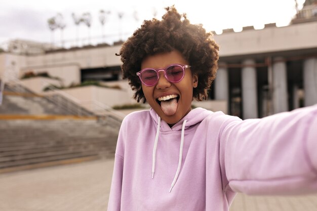 La joven y alegre mujer rizada con coloridas gafas de sol rosas y una sudadera con capucha morada hace una cara divertida, muestra la lengua y se toma una selfie afuera