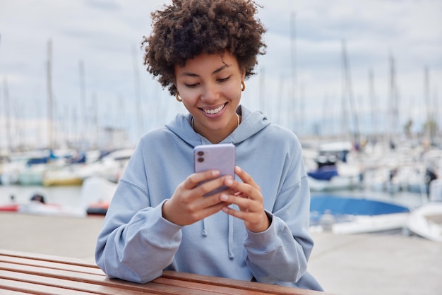 Joven alegre mujer rizada usa teléfono móvil se sienta en el muelle cerca del mar lee un mensaje de texto agradable usa sudadera con capucha se relaja al aire libre pasa tiempo libre en el puerto marítimo estando de buen humor Concepto de tecnología