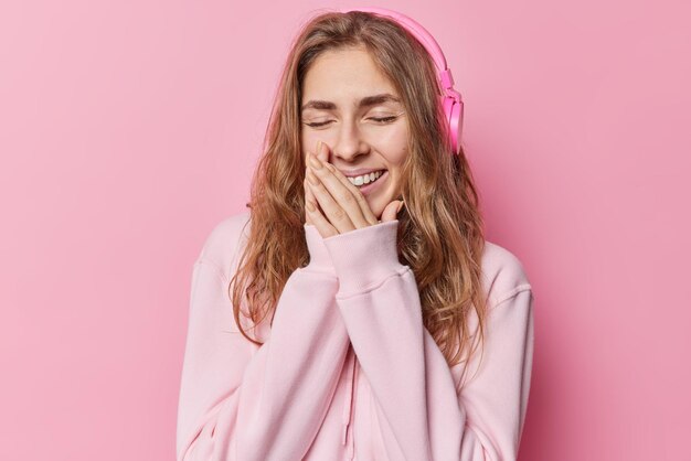 Una joven alegre de pelo largo se ríe positivamente y mantiene las manos cerca de la boca escucha su música favorita a través de auriculares vestida con una cómoda sudadera que mantiene los ojos cerrados aislados sobre un fondo rosa
