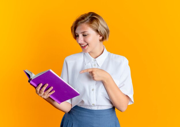 Joven alegre rubia rusa mira y apunta a libro aislado sobre fondo naranja con espacio de copia