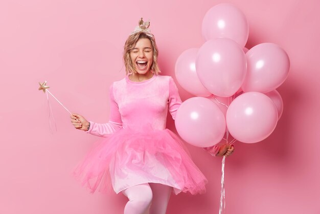 Una joven alegre y sincera con el peinado exclama de la felicidad que usa corona y el vestido sostiene un montón de globos inflados con una varita mágica que disfruta de la celebración y el tiempo de fiesta aislado sobre un fondo rosa.