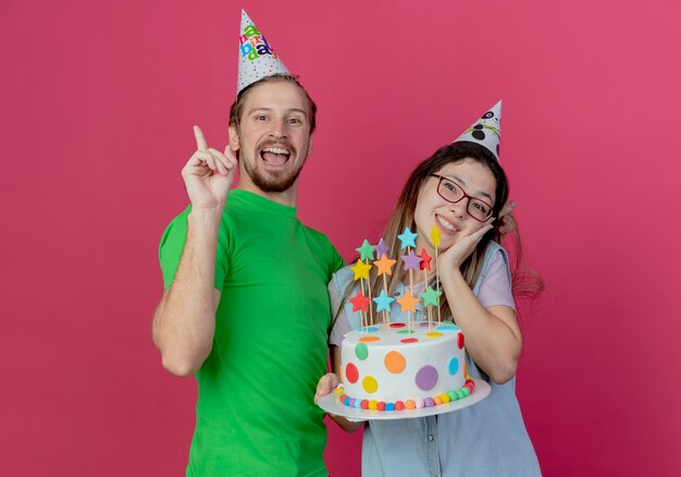 Joven alegre con sombrero de fiesta apunta hacia arriba y se encuentra con una niña feliz con sombrero de fiesta sosteniendo pastel de cumpleaños aislado en la pared rosa