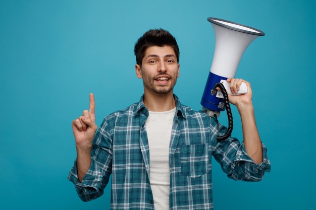 Un joven alegre sosteniendo un altavoz mirando a la cámara apuntando hacia arriba aislado de fondo azul