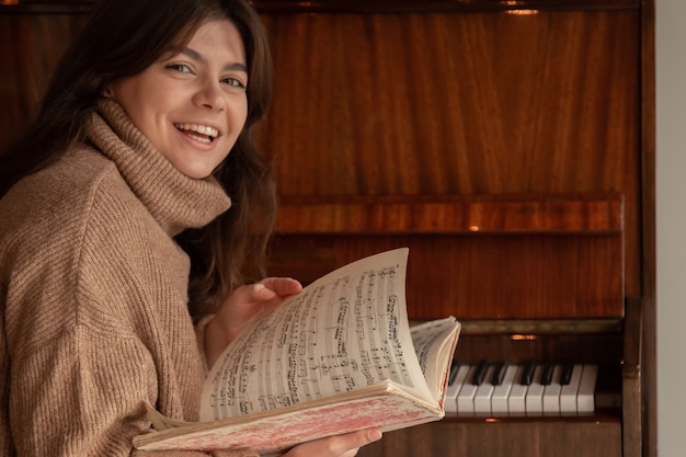 Una joven alegre con un suéter acogedor se sienta cerca del piano y mira las notas.