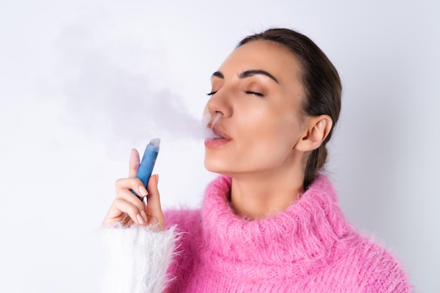 Foto gratuita una joven alegre con un suéter de colores brillantes en un fondo blanco disfruta de un cigarrillo electrónico desechable con los ojos cerrados fuma libera humo