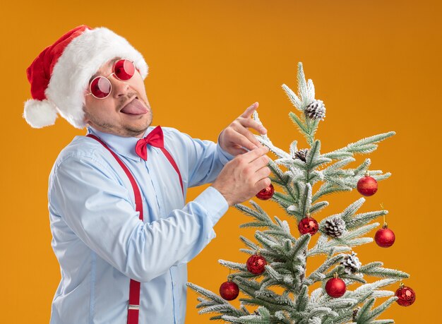 Joven alegre con tirantes pajarita con gorro de Papá Noel y gafas rojas de pie cerca del árbol de Navidad que cuelga el juguete en el árbol feliz y sorprendido sacando la lengua sobre fondo naranja