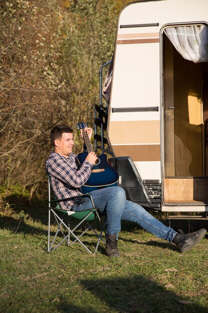 Joven alegre tocando su guitarra frente a su autocaravana retro en las montañas. Hombre relajante en las montañas.