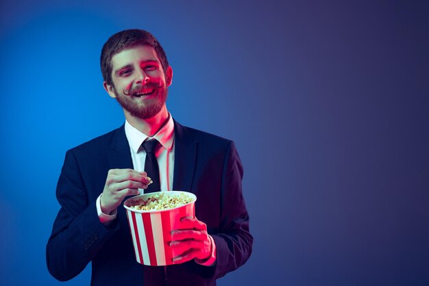 Joven alegre con traje oficial comiendo palomitas de maíz viendo películas aisladas sobre el fondo azul del estudio con luz de neón