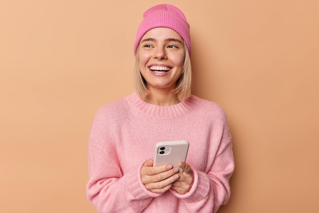 Foto gratuita una joven alegre usa el teléfono móvil y envía mensajes de texto. mira hacia otro lado. tiene una amplia sonrisa. pide algo en la tienda en línea. lleva un puente rosa y un sombrero aislado sobre un fondo marrón. concepto de tecnología.