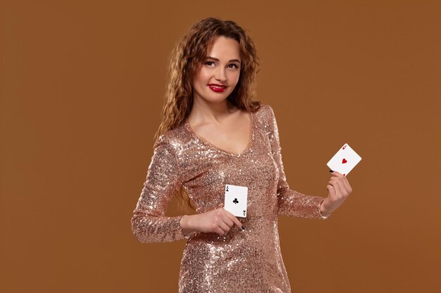 Una joven alegre, vestida con un vestido dorado brillante, sostiene dos cartas en las manos, sonríe y mira la cámara. Concepto de casino sobre un fondo marrón.