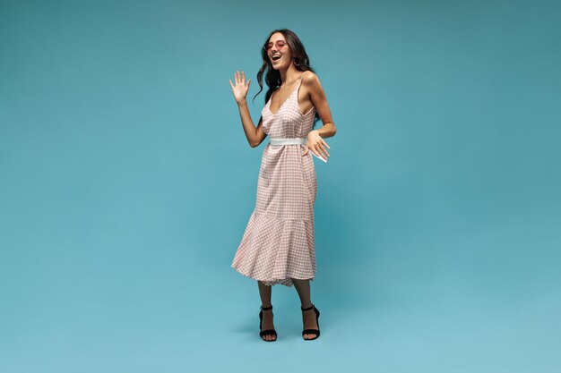 Una joven alegre con un vestido midi de gafas de sol rosas brillantes y tacones negros bailando y riendo en un fondo azul aislado