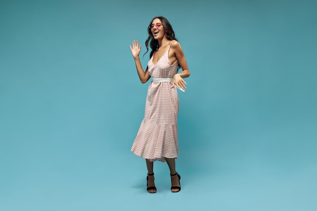 Una joven alegre con un vestido midi de gafas de sol rosas brillantes y tacones negros bailando y riendo en un fondo azul aislado