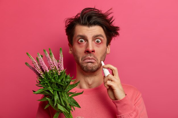 Un joven alérgico enfermo gotea la nariz con gotas nasales, tiene los ojos y la nariz enrojecidos, alergia a la planta, síntomas de rinitis o fiebre del heno, mira fijamente, posa contra la pared rosada, reacciona con el polen