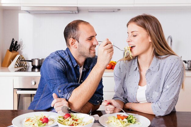 Joven alimentación de ensalada a su esposa sentada en la cocina
