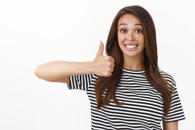 Una joven amable y comprensiva que sonríe felizmente aprueba una buena elección mostrando el pulgar hacia arriba en un signo de aprobación y una sonrisa de aceptación y asiente con la cabeza en respuesta afirmativa de pie con fondo blanco en una camiseta a rayas