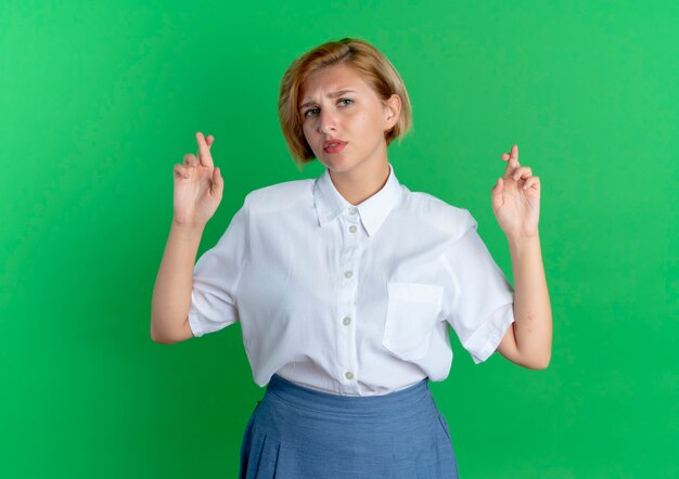 Joven ansiosa rubia rusa cruza los dedos aislados sobre fondo verde con espacio de copia