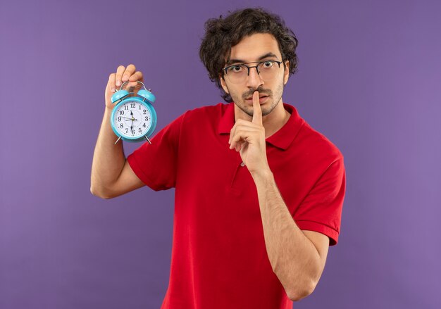 Joven ansioso en camisa roja con gafas ópticas tiene reloj y gestos signo de silencio aislado en la pared violeta