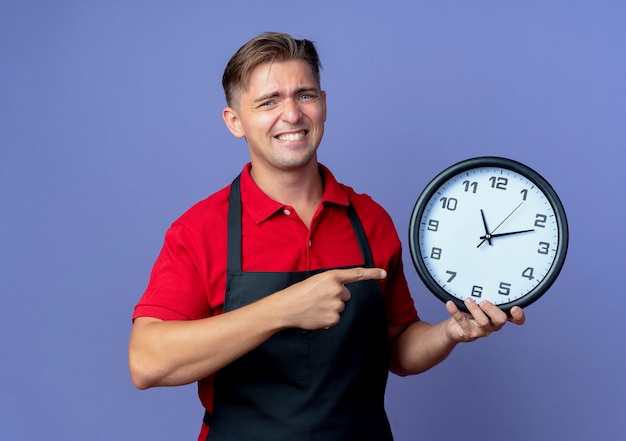 Joven, ansioso, rubio, macho, peluquero, en, uniforme, sostiene, y, puntos, en, reloj, aislado, en, violeta, espacio, con, espacio de copia