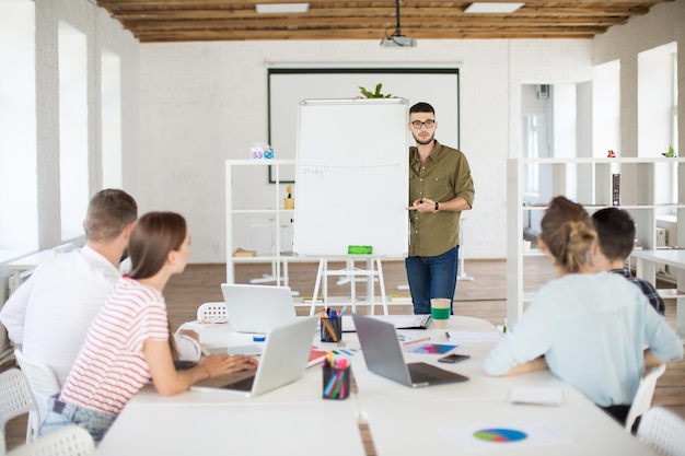 Joven con anteojos y camisa de pie cerca de una pizarra blanca mientras discute cuidadosamente un nuevo proyecto con colegas Grupo de personas creativas que trabajan juntas en una oficina blanca moderna