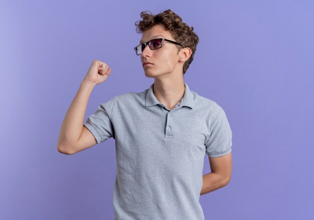 Joven de anteojos negros vistiendo camisa polo gris mirando a un lado con la cara de serioua apuntando hacia atrás sobre la pared azul