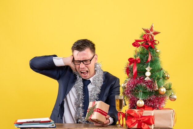 Joven con anteojos sosteniendo su oreja sentado en la mesa cerca del árbol de Navidad y presenta en amarillo