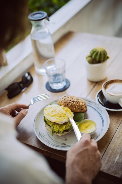 Joven apuesto con una camisa blanca abierta, desayunando en un café con una hamburguesa vegetariana, bebiendo café, estilo de vida en una isla tropical, vida en Bali.