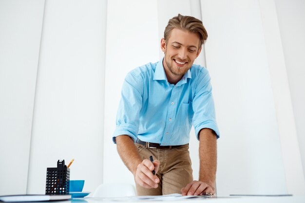 Joven apuesto confiado alegre sonriente hombre de negocios trabajando de pie en la mesa de dibujo boceto. Interior de oficina moderno blanco