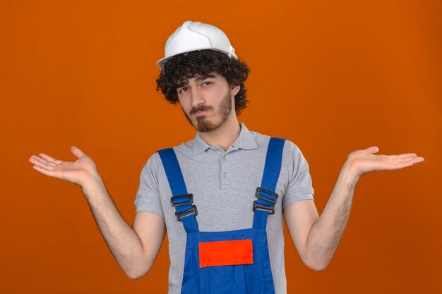 Joven apuesto constructor barbudo vistiendo uniforme de construcción y casco de seguridad despistado y confundido con los brazos abiertos sin idea de concepto de pie sobre la pared naranja aislada