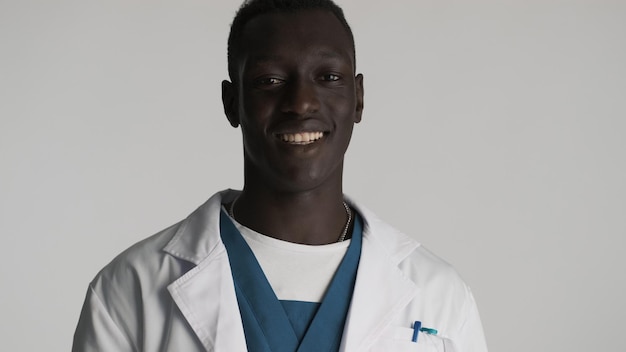Joven y apuesto doctor afroamericano que parece feliz sonriendo a la cámara sobre fondo blanco