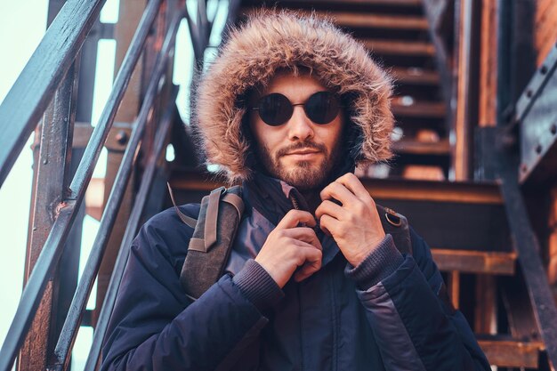 Un joven apuesto y elegante con un abrigo de invierno y gafas de sol sentado en las escaleras afuera.