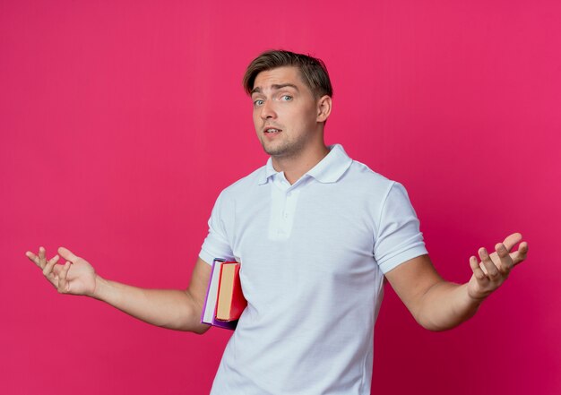 Joven apuesto estudiante sosteniendo libros y extiende las manos aisladas en la pared rosa