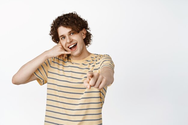 Un joven apuesto y feliz señalando con el dedo a la cámara mostrando un gesto de llamada de teléfono móvil invitándote a pararte sobre fondo blanco