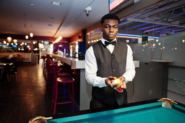 Joven apuesto hombre africano vestido con camisa blanca, chaleco negro y corbata de moño, jugar al billar