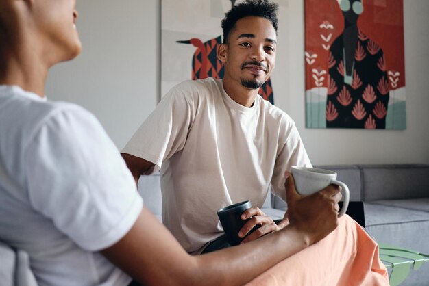 Joven apuesto hombre afroamericano casual mirando a la cámara durante un descanso para tomar café con un amigo en un moderno espacio de trabajo conjunto