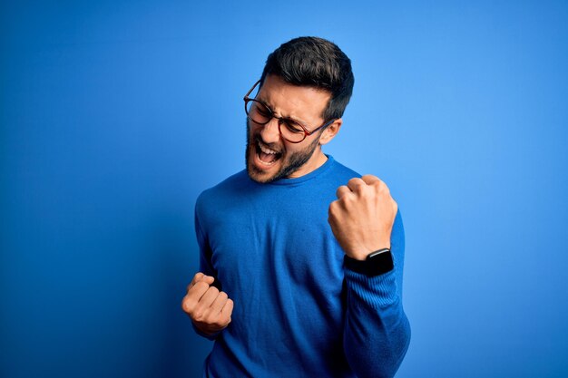 Joven apuesto hombre con barba usando suéter casual y anteojos sobre fondo azul celebrando sorprendido y asombrado por el éxito con los brazos levantados y los ojos cerrados Concepto de ganador