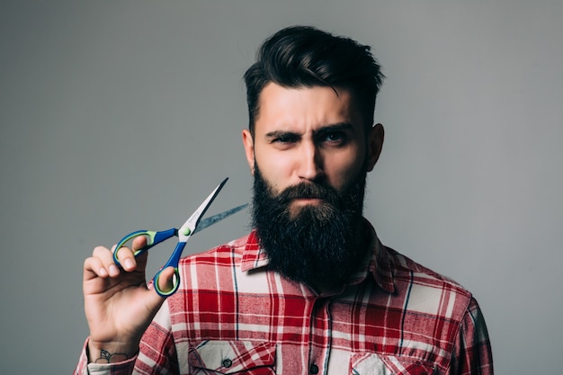 Joven apuesto hombre barbudo con bigote de barba larga y cabello morena con tijeras de peluquero o peluquero con cara emocional en pared gris